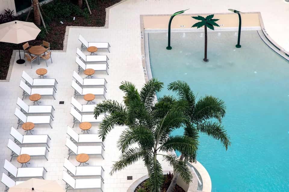 Aerial view of resort pool with turquoise water, loungers with tables, palm trees, and white patio area