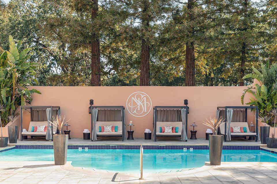 Luxury resort pool with cabanas and pink wall backdrop surrounded by tall trees at sunset