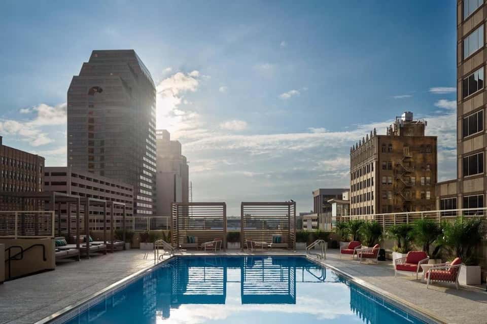 Rooftop pool deck with city skyline, lounge chairs, umbrellas, and palm trees in urban setting
