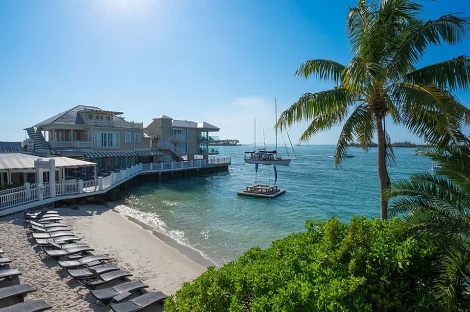 Tropical beachfront resort with stilted buildings, palm trees, sailboats, and sandy beach with loungers