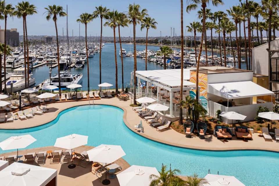 Aerial view of resort pool deck with palm trees, lounge chairs, and marina with sailboats