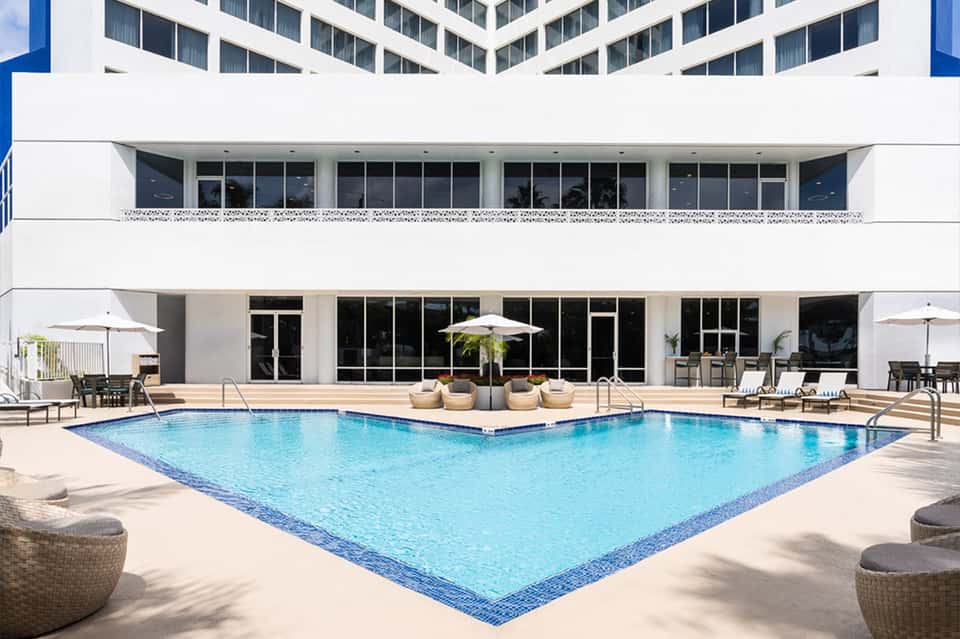 Large rectangular resort pool with lounge chairs, white umbrellas, modern building facade, and seating areas