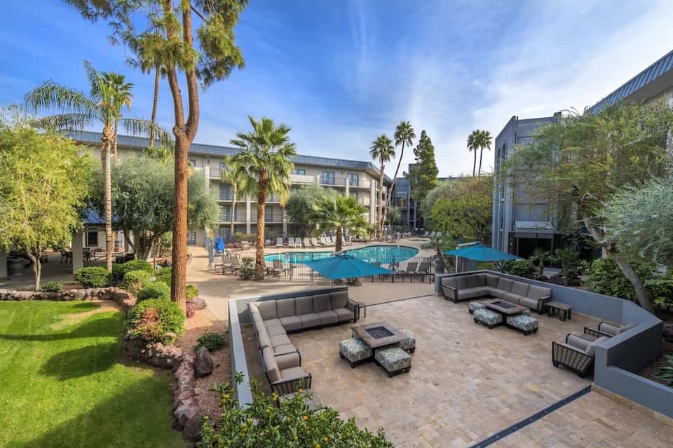 Resort courtyard with curved seating areas, fire pit, pool, palm trees, and modern buildings