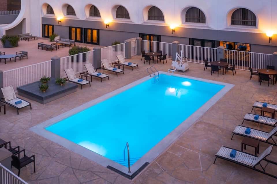 Illuminated resort pool deck at dusk with lounge chairs, white railings, and arched windows of the main building