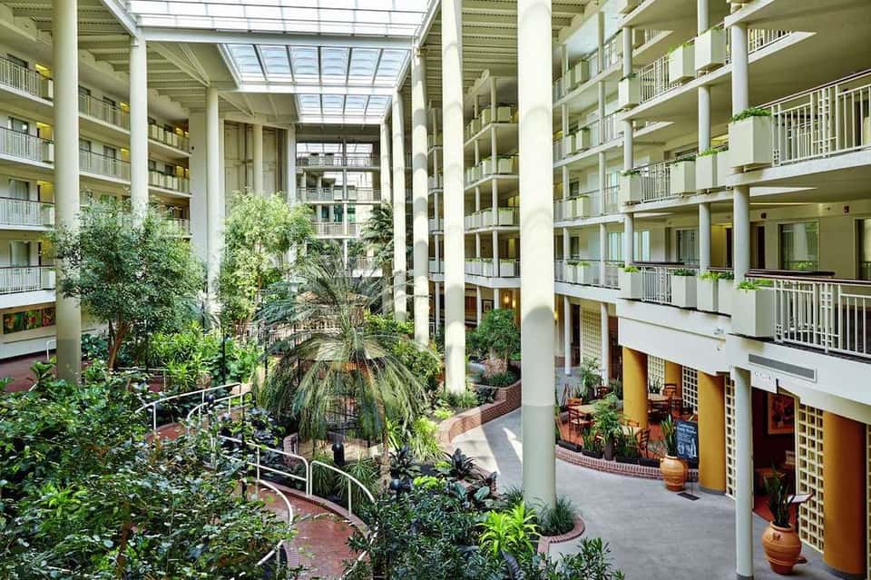 Modern hotel atrium with multi-level balconies, indoor plants, skylights, and brick flooring