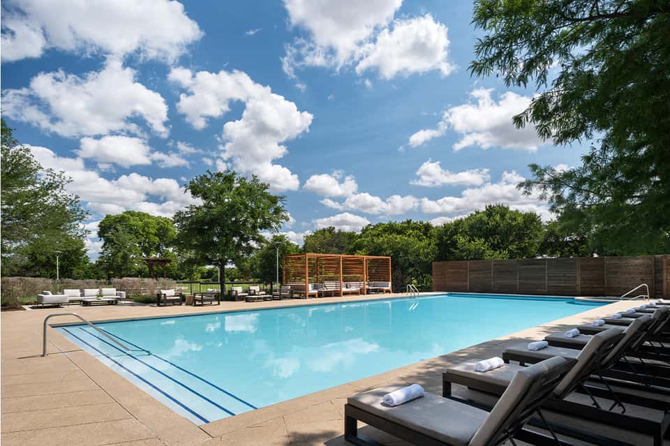 Resort pool surrounded by lounge chairs, wooden pergolas, and lush green landscaping under blue sky