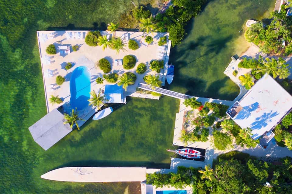 Aerial view of waterfront resort with blue pool, palm trees, docked boats, and surrounding green landscape.