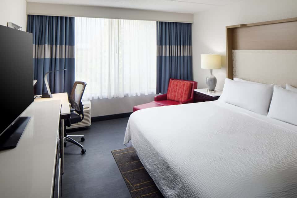 Modern hotel bedroom with king bed, work desk, blue striped curtains, and city view windows