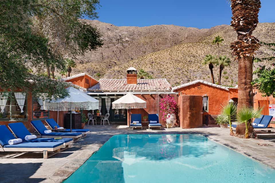 Desert resort pool with terracotta buildings, palm trees, and lounge chairs under umbrellas