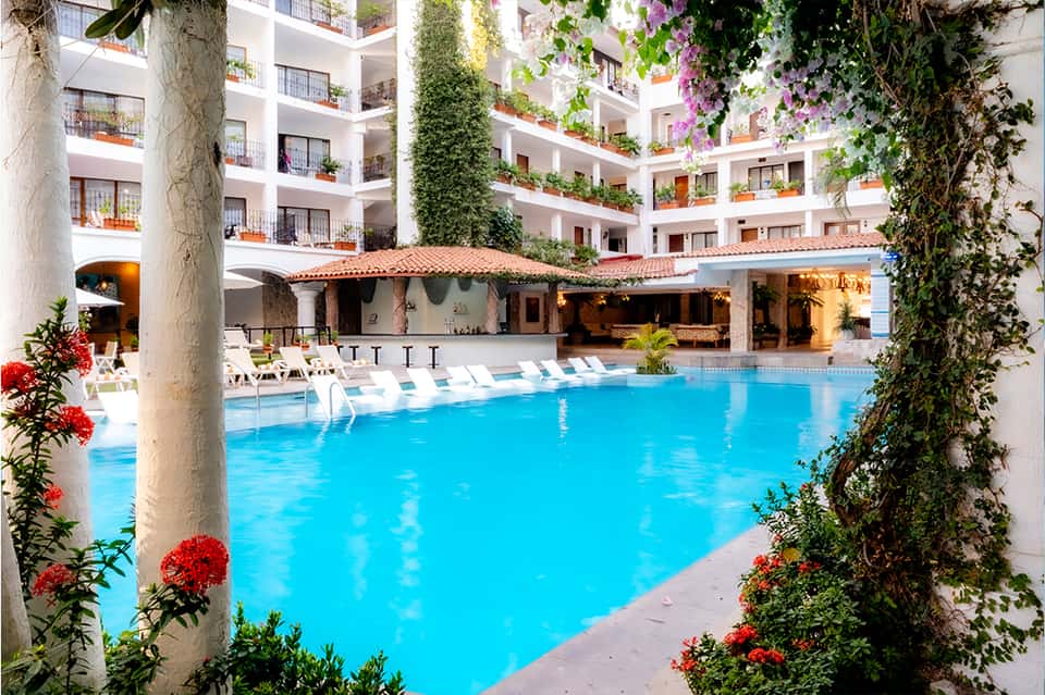 Bright blue resort pool surrounded by white loungers, ivy-covered columns, and multi-story white buildings