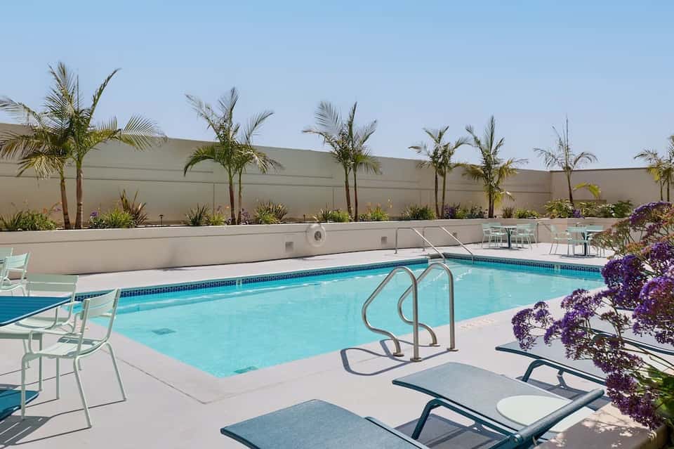 Rooftop pool with turquoise water, lounge chairs, palm trees, and high concrete walls under clear sky