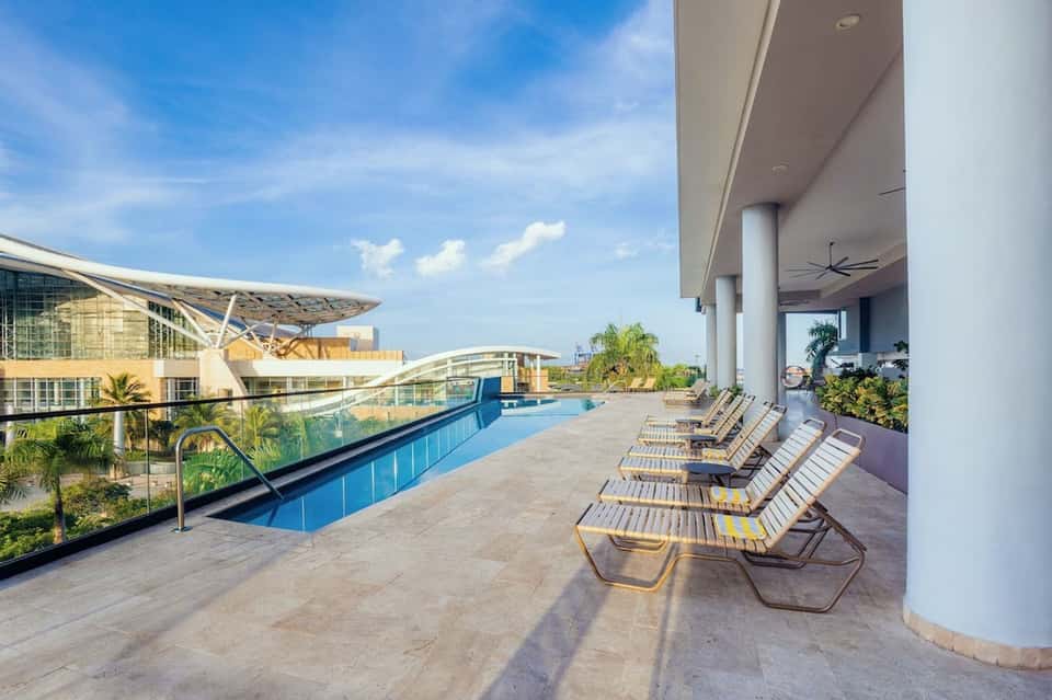 Modern resort pool deck with lounge chairs, architectural building, palm trees, and blue water