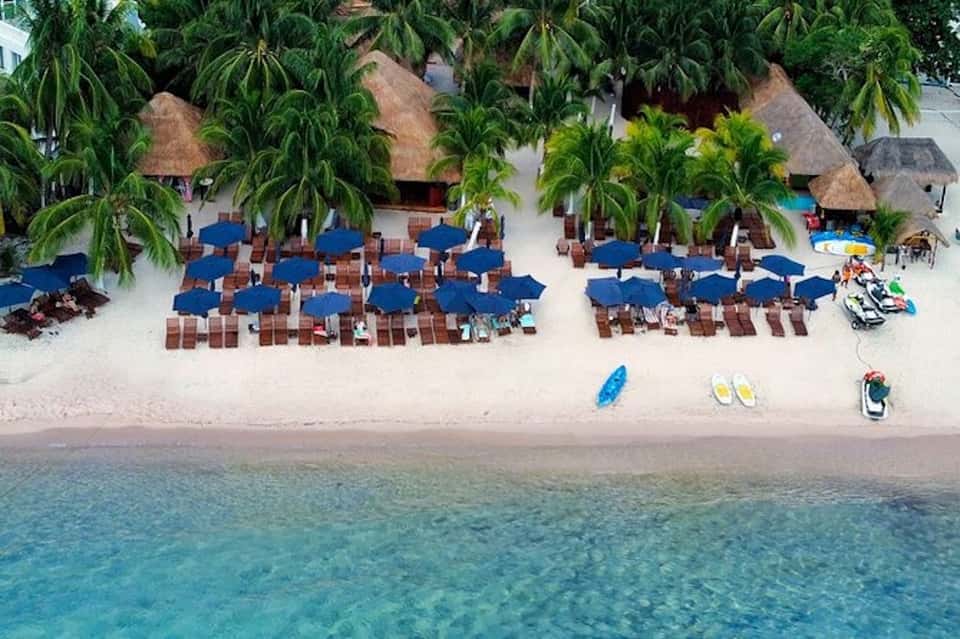 Aerial view of tropical beach resort with blue umbrellas, thatched-roof bungalows, and palm trees