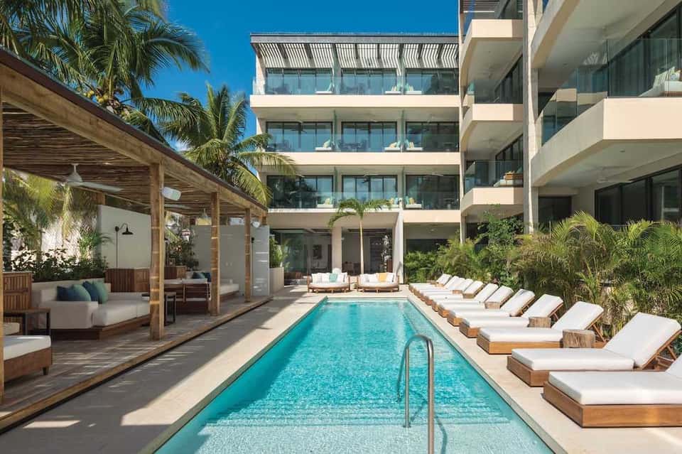 Contemporary resort pool with wooden lounge chairs, cabana, and multi-story building backdrop