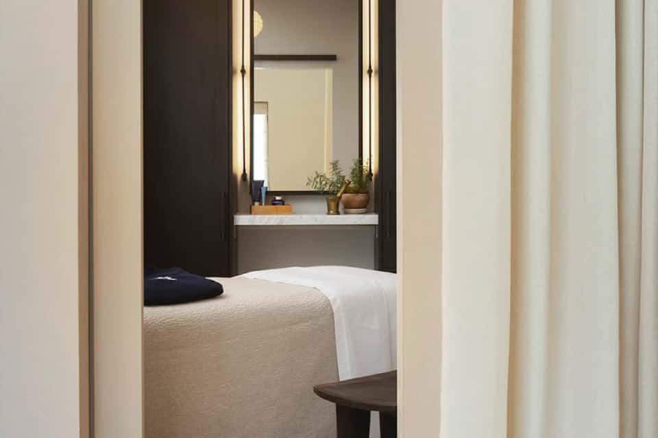 Minimalist hotel room with bed, illuminated mirror, vanity area, and neutral color palette viewed through doorway