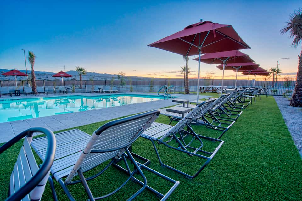 Resort pool at dusk with rows of lounge chairs under burgundy umbrellas and palm trees
