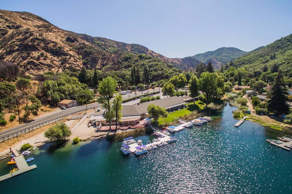Aerial view of lakeside resort nestled in green mountains with boats, buildings, and cypress trees