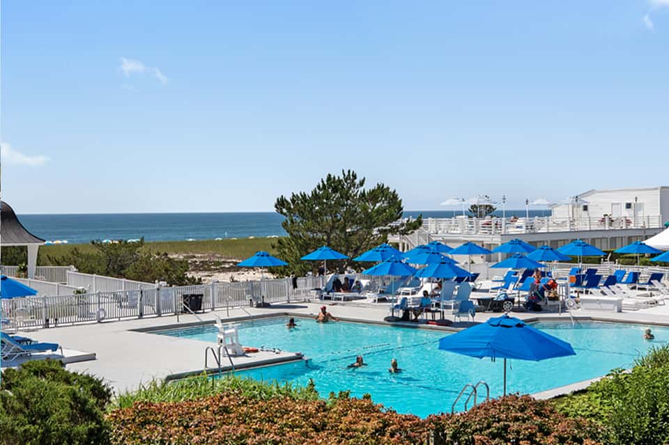 Resort pool area with bright blue umbrellas, loungers, ocean view, and white buildings in background
