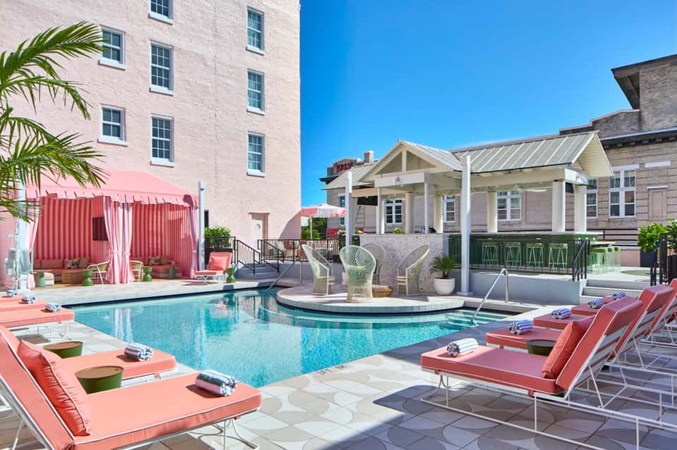 Resort pool area with pink lounge chairs, white pergola, palm trees, and multi-story hotel building