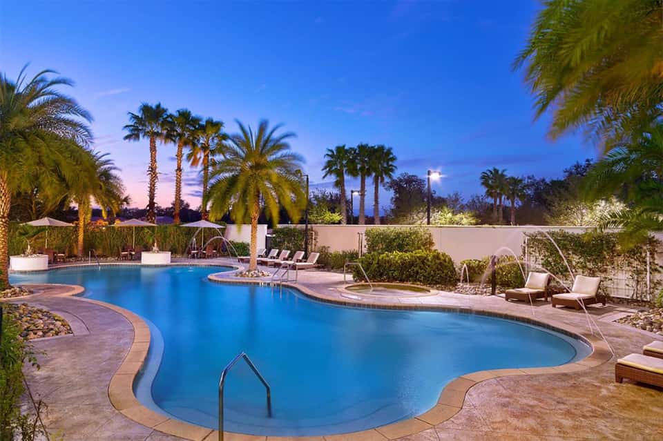 Resort pool at dusk surrounded by palm trees, lounge chairs, and white walls under blue sky