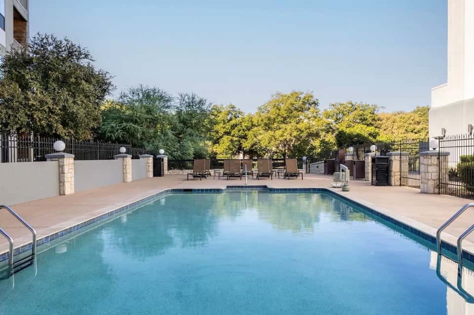 Resort pool with blue water, white concrete deck, lounge chairs, and landscape surrounding fence