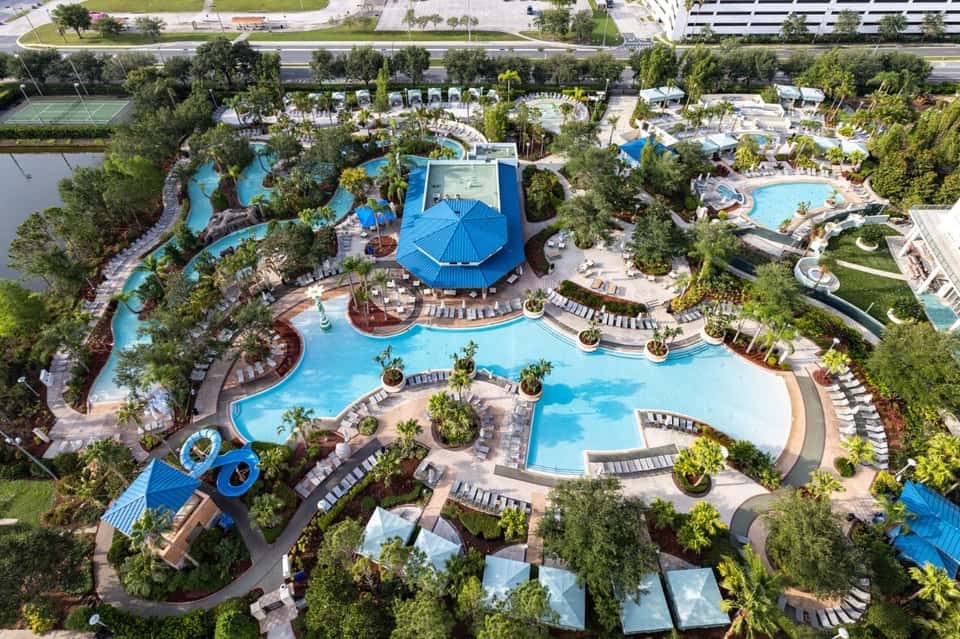 Aerial view of sprawling resort with multiple pools, blue umbrellas, water features, and landscaping