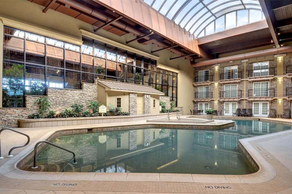 Indoor heated swimming pool with stone walls, arched glass ceiling, and multi-story hotel rooms