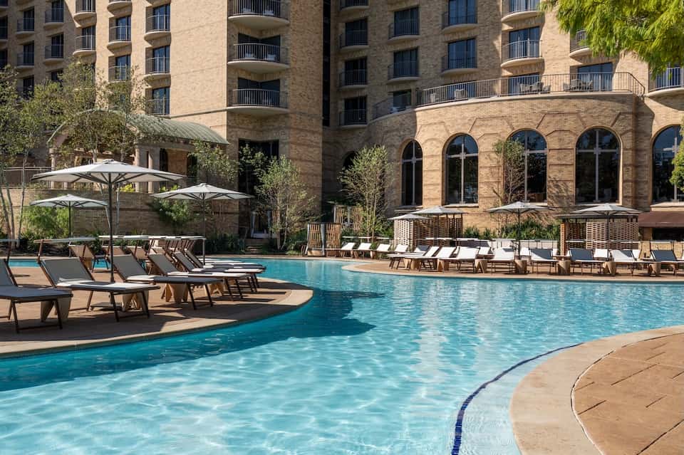 Curved outdoor pool at brick hotel with lounge chairs, umbrellas, and multi-story residential building