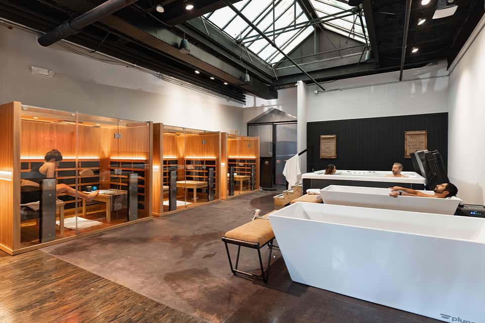 Spa facility with wooden sauna cabins on left and white soaking tubs with guests on right under industrial ceiling