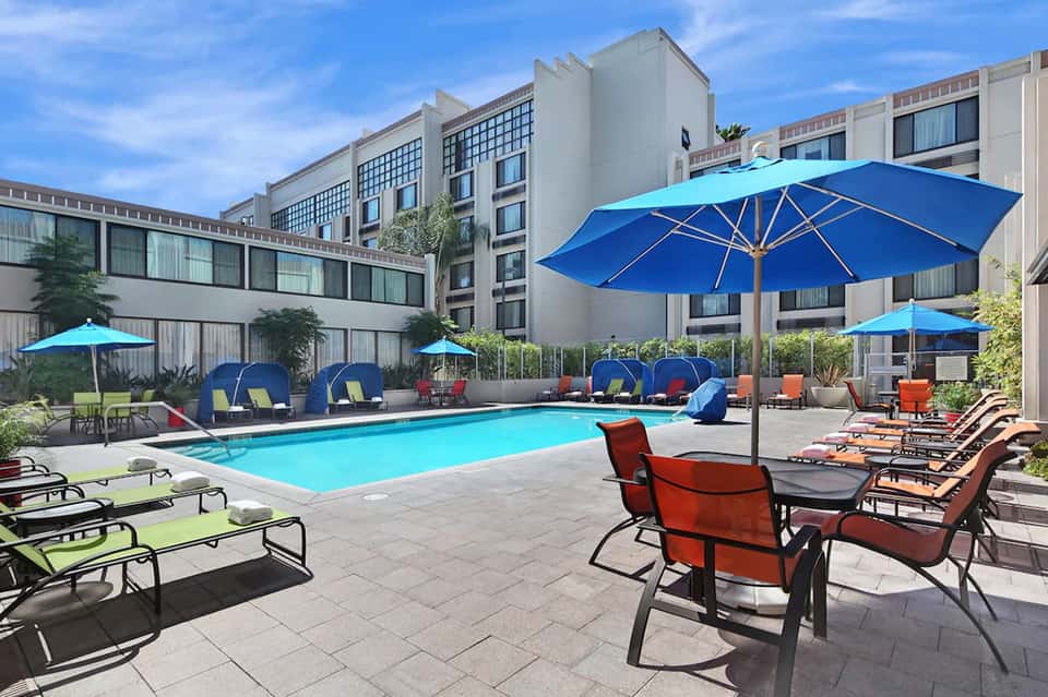 Resort pool area with blue umbrellas, colorful lounge chairs, and multi-story building in background