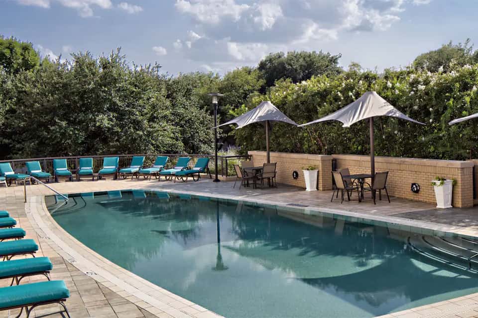 Curved resort pool with turquoise loungers, white sail umbrellas, and brick building with natural backdrop