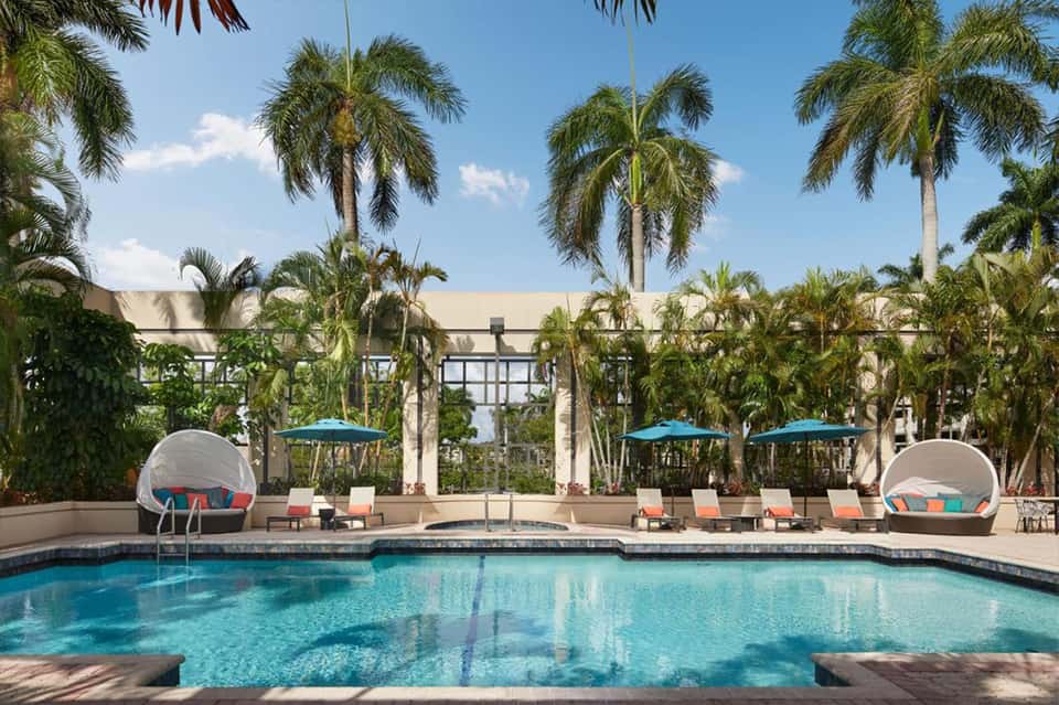 Resort pool with turquoise water, palm trees, lounge chairs, umbrellas, and modern architecture