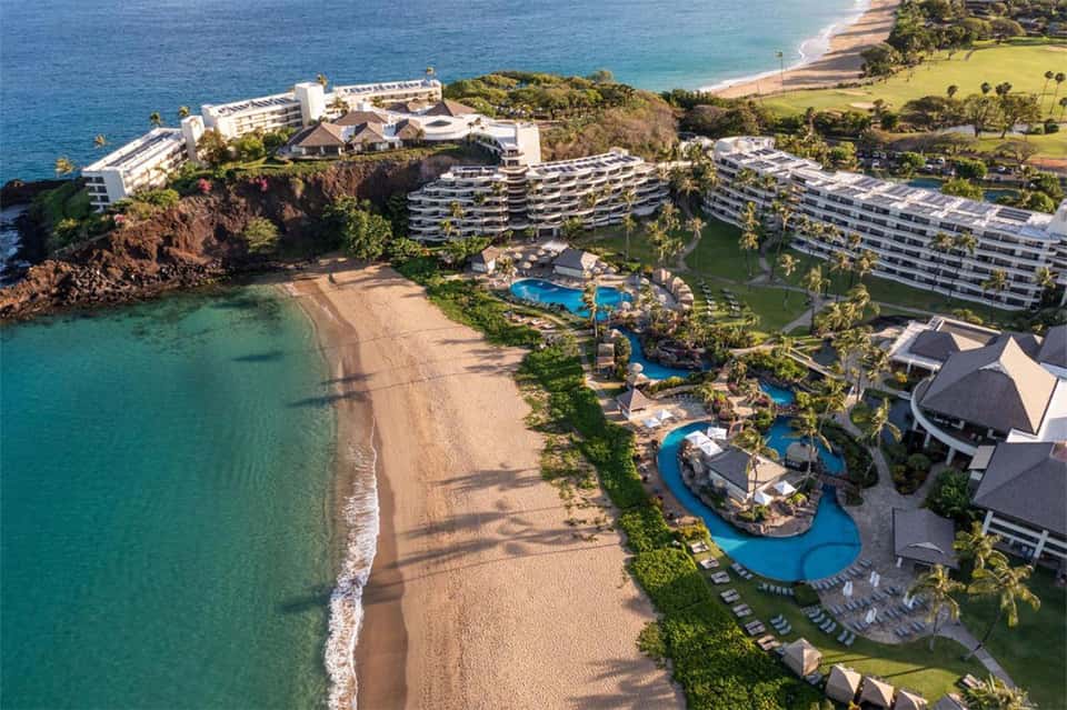 Aerial view of beachfront resort with curved pools, palm trees, buildings, and pristine turquoise ocean