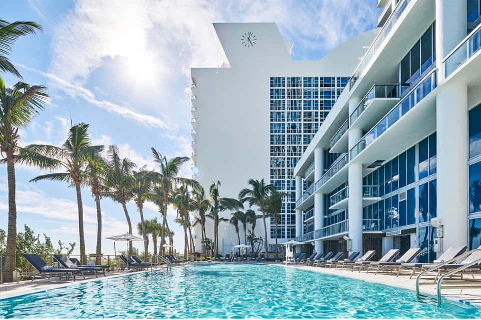 Large resort pool with turquoise water, lounge chairs, palm trees, and modern white building