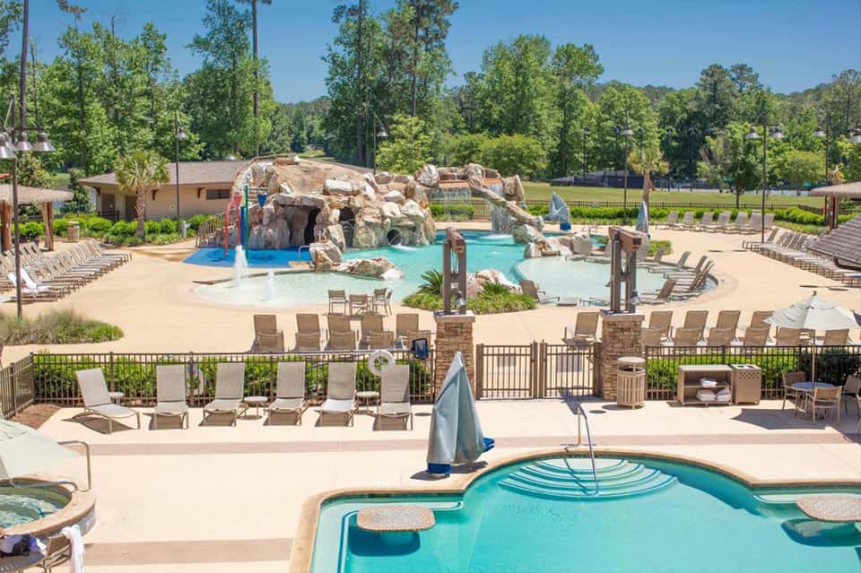 Expansive resort pool area with water feature, rock formation, multiple pools, lounge chairs, and wooded surroundings