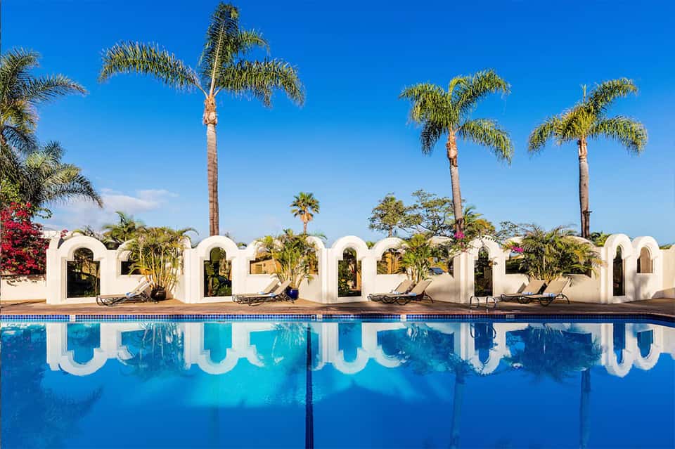 Resort pool with white arched cabanas, palm trees, red flowers, and clear blue water under sunny sky
