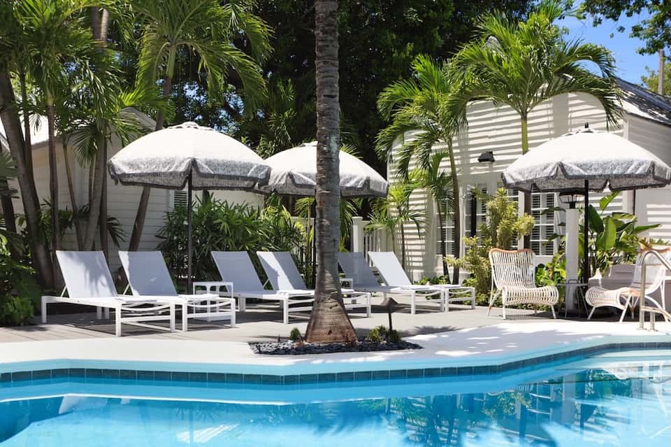 Resort pool with white lounge chairs, thatched umbrellas, palm trees, and beachfront cabins