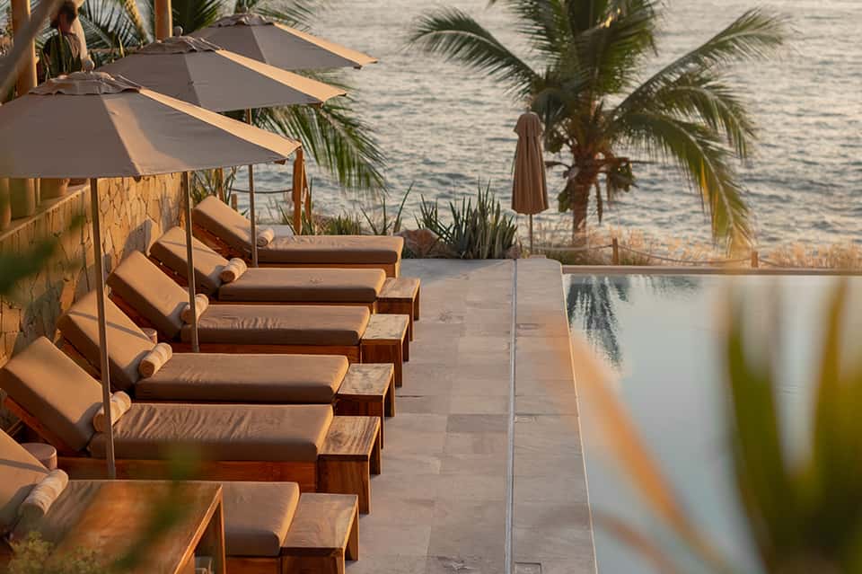 Beachfront lounge area with cushioned sunbeds under umbrellas, palm trees, and ocean view at golden hour