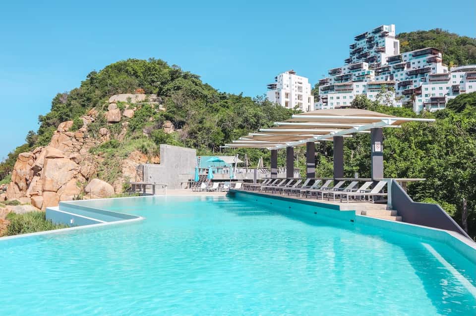 Stunning infinity pool overlooking rocky hillside with residential buildings, loungers, and pergola shade
