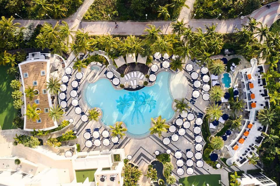 Aerial view of large resort pool complex with multiple umbrellas and palm trees surrounded by buildings
