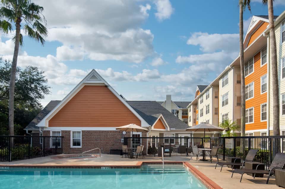 Resort pool with brick clubhouse, lounge chairs, palm trees, and colorful residential buildings