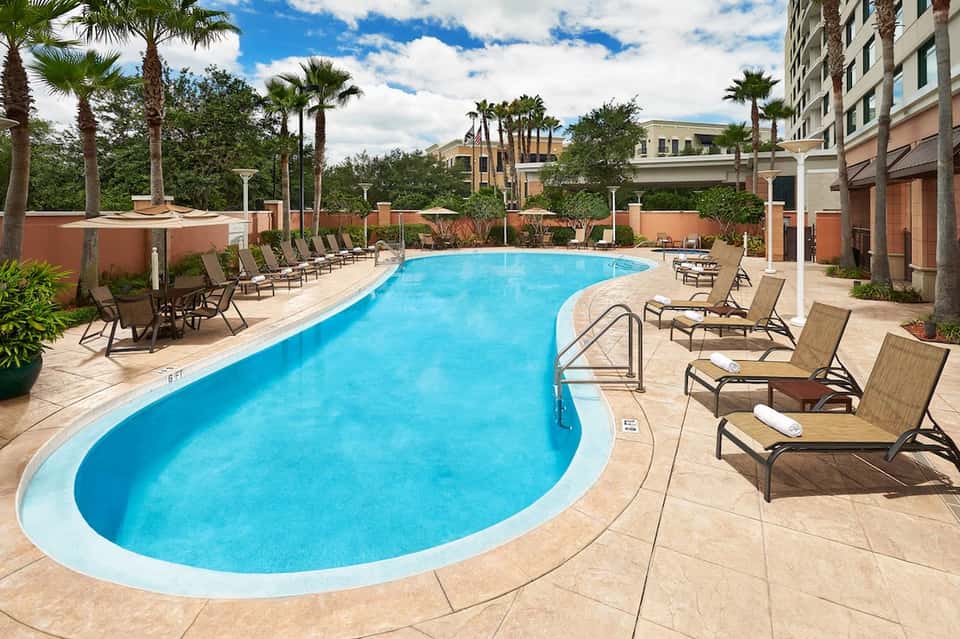 Resort pool with lounge chairs, palm trees, stone patio, and multi-story hotel building backdrop