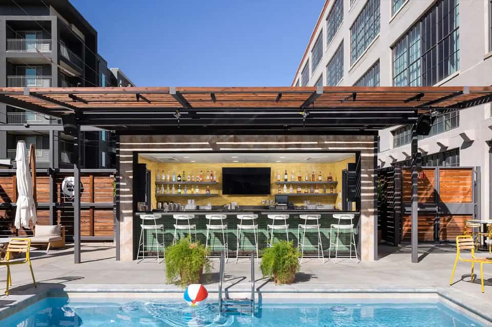 Modern rooftop pool bar with yellow backlit shelving, white bar seating, and pergola
