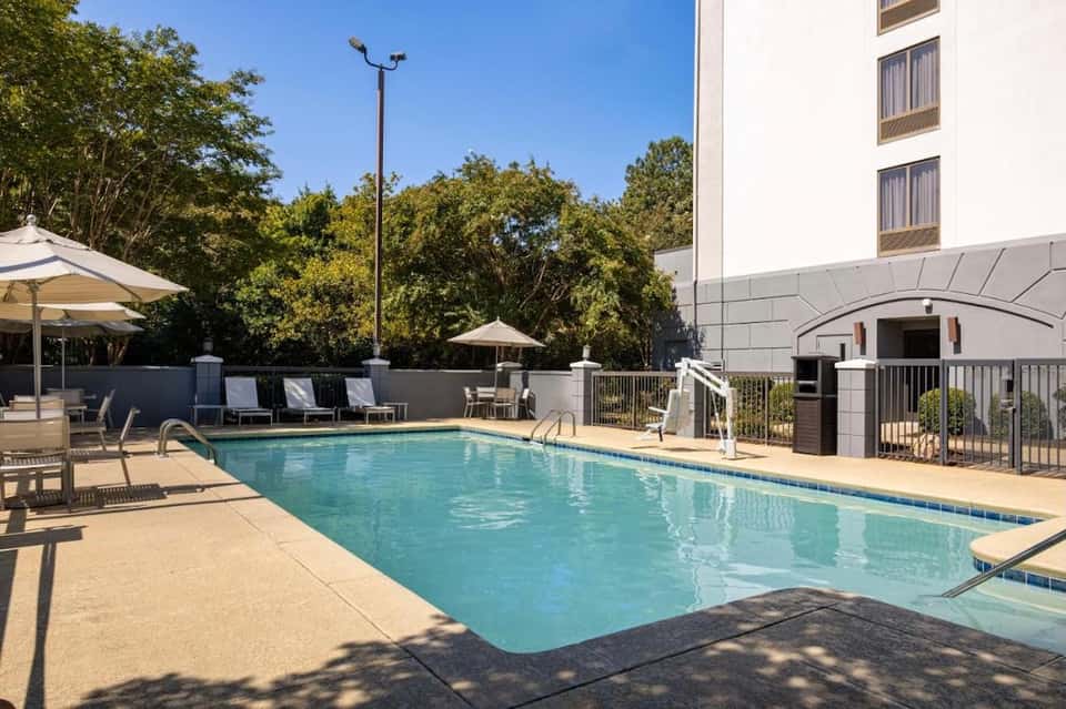 Hotel pool surrounded by lounge chairs, umbrellas, trees, and multi-story building with railing