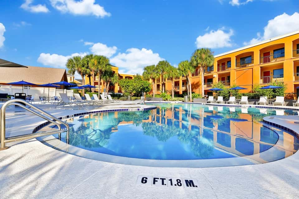 Resort pool with depth marking, surrounded by yellow buildings, palm trees, and lounge seating areas