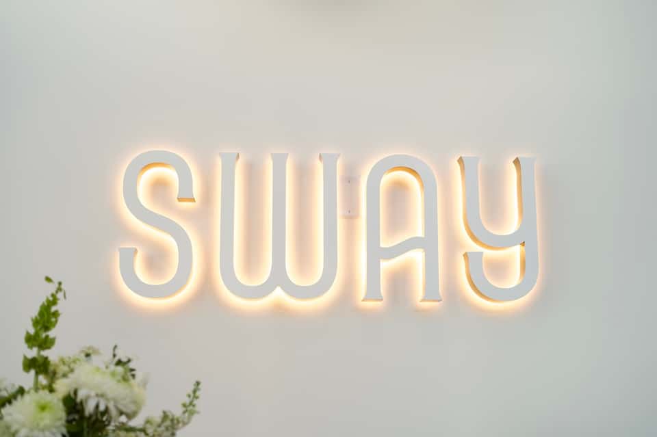 SWAY neon sign glowing warmly on white wall beside flowers