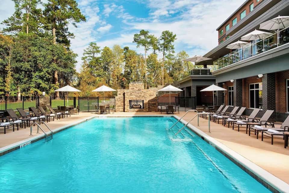 Resort swimming pool with lounge chairs, white umbrellas, scenic forest backdrop, and brick building