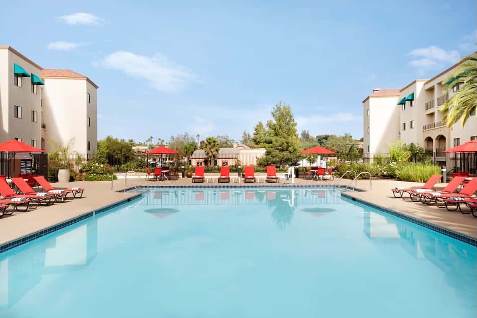 Resort swimming pool with red loungers and umbrellas, surrounded by apartment buildings and landscaping