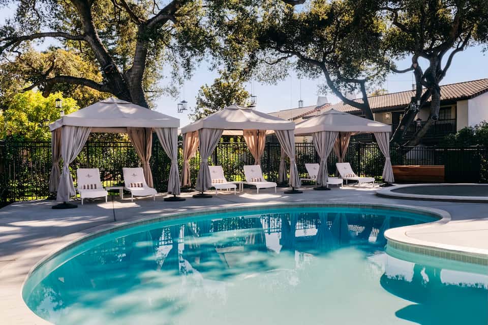 Resort pool with lounge chairs under beige pergolas, large oak trees providing shade, spa building in background
