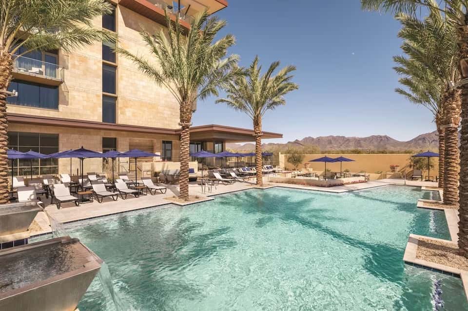 Desert resort pool with turquoise water, palm trees, lounge chairs, and mountain views